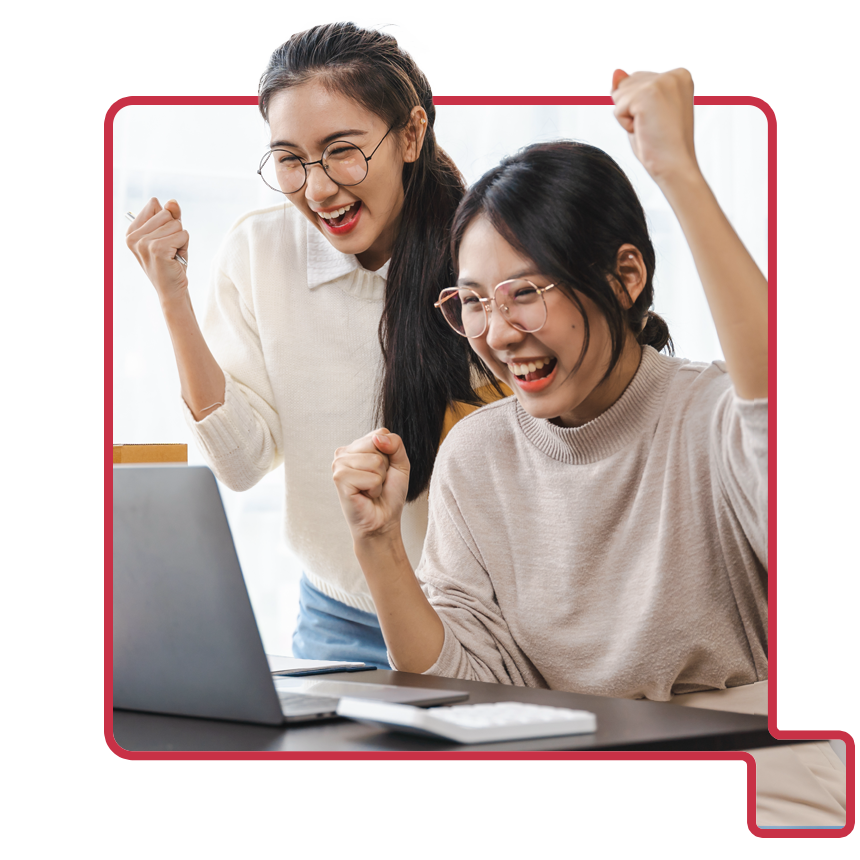 Two young women celebrating with raised fists while looking excitedly at a laptop screen in a bright office setting.